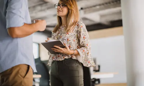 Smilende kollega som holder et nettbrett mens hun snakker med en medarbeider i et moderne kontormiljø, og fremmer samarbeid og teamarbeid
